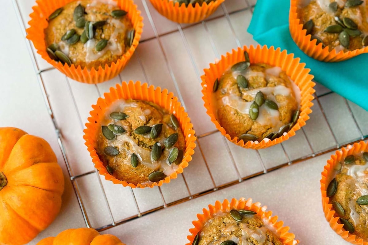 Pumpkin muffins with glaze and seeds in orange liners on a cooling rack with pumpkins