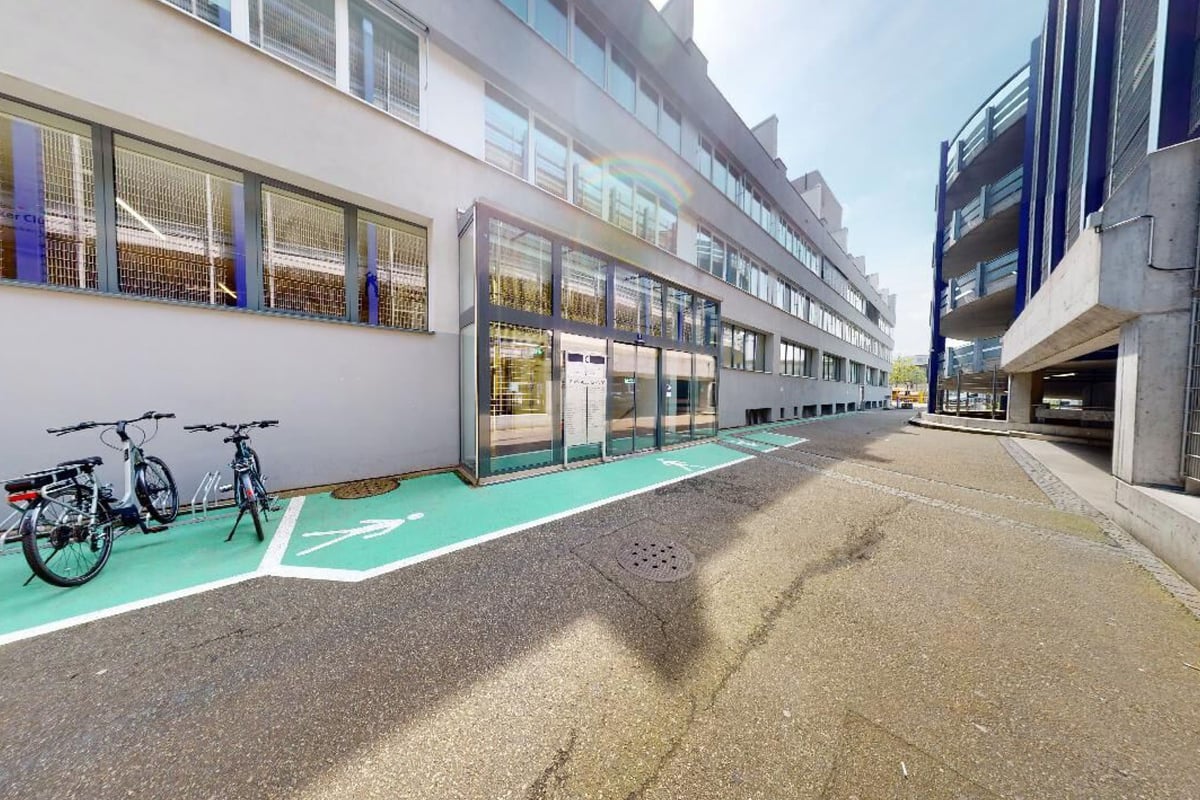 PureGym Basel Dreispitz: Exterior alley with green bike lane, parked bicycles near building storefronts.
