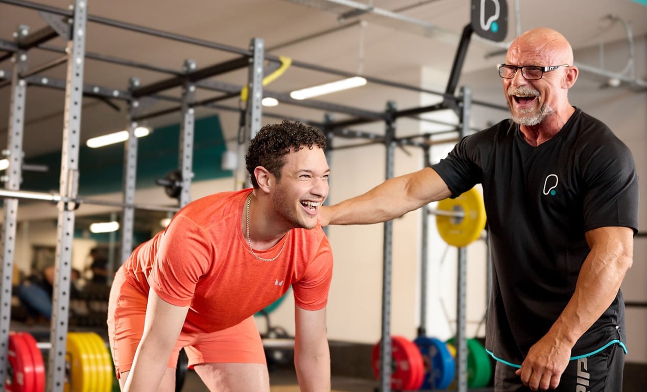 PureGym: a smiling male customer performing a deadlift being encouraged by a male personal trainer