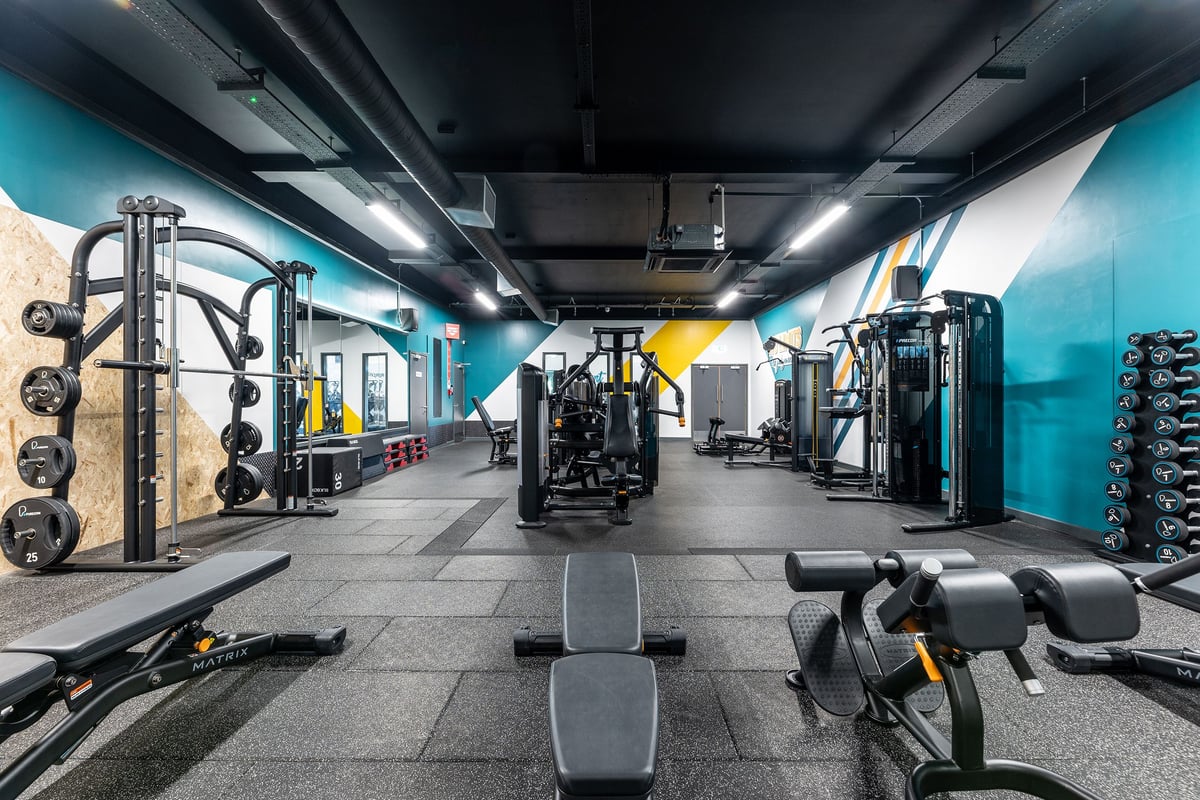 PureGym Leamington Spa: Weights racks, cable machines, benches, rubberized floors in a teal and yellow-accented gym studio.