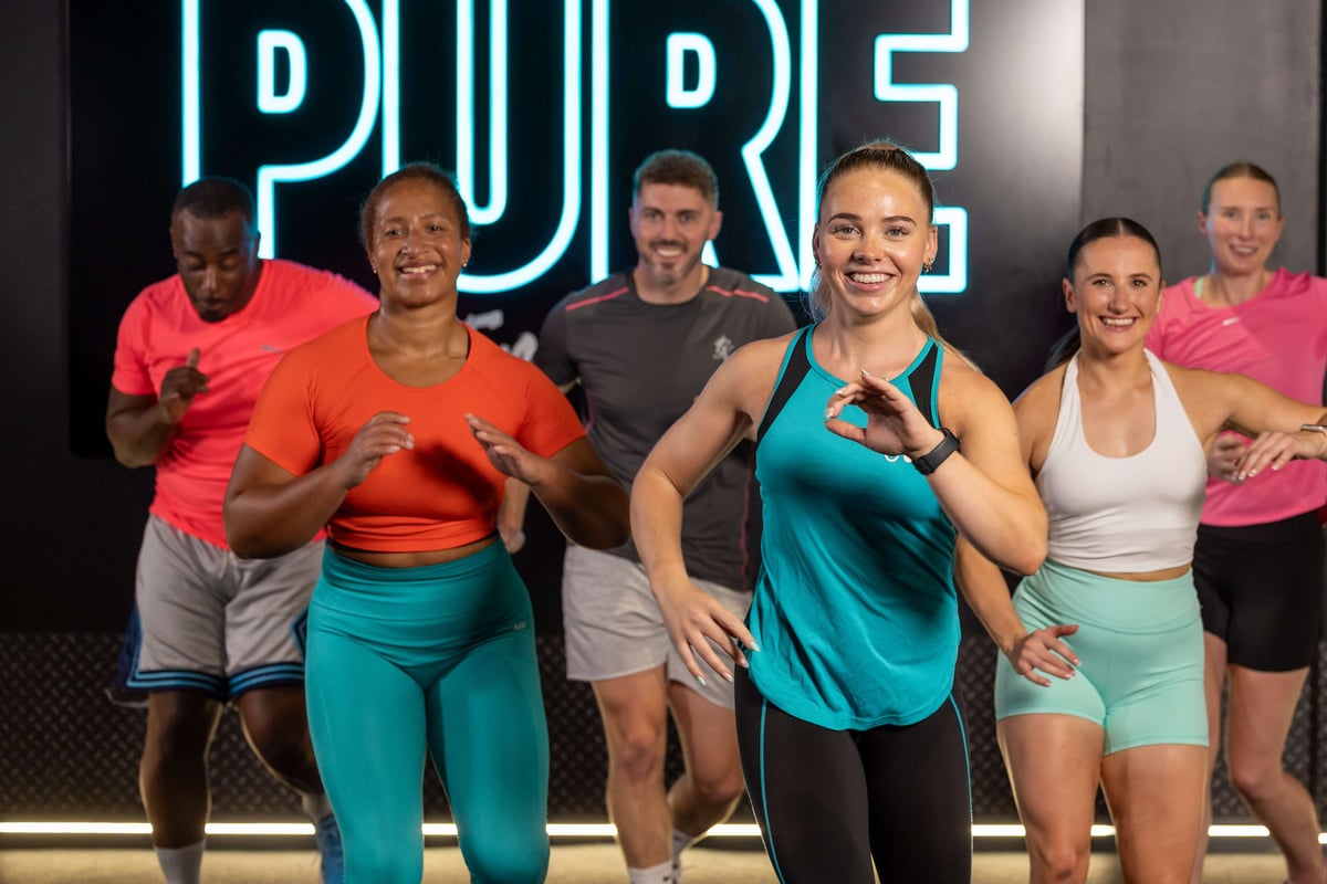 Group of six people smiling and exercising indoors with a neon PURE sign behind them