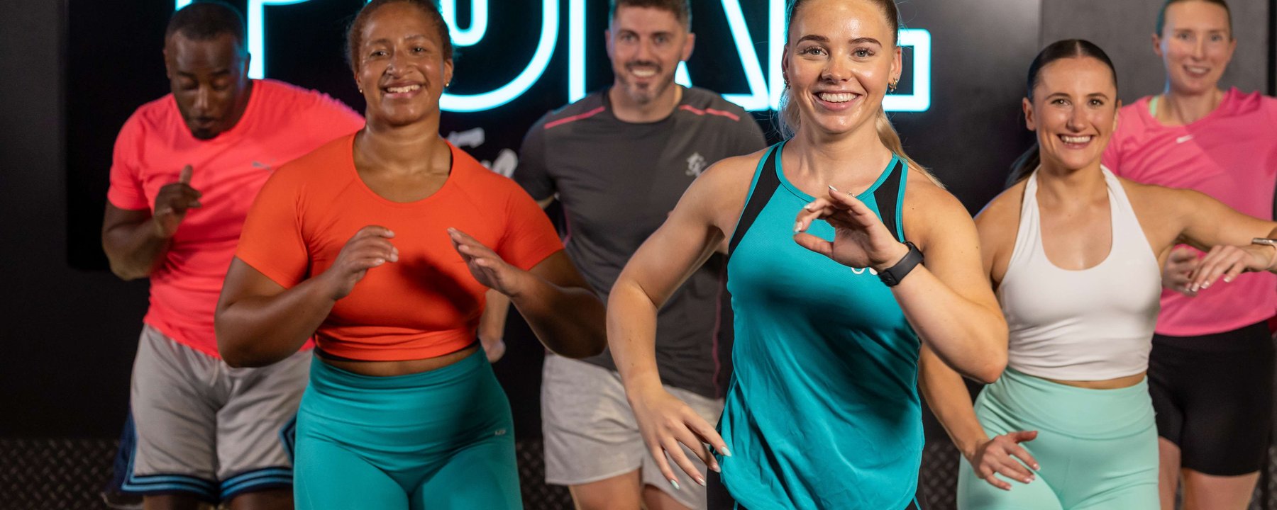 Group of six people smiling and exercising indoors with a neon PURE sign behind them