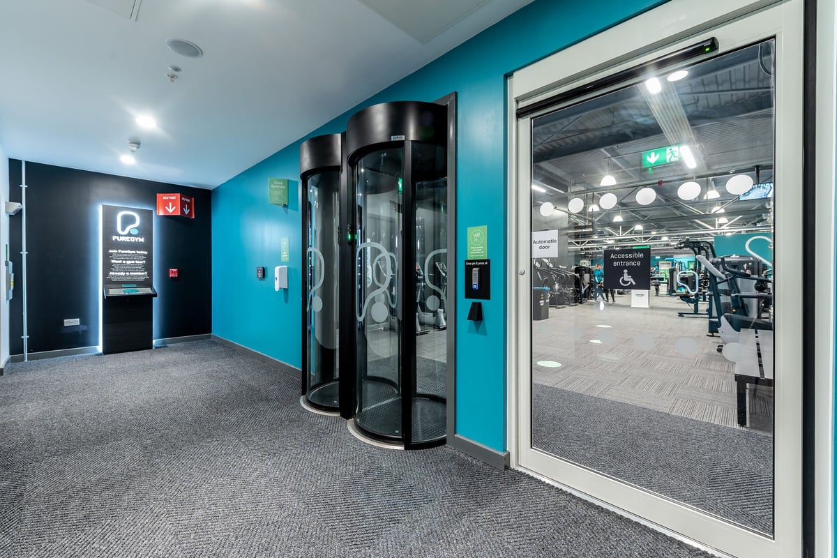 PureGym Reading Calcot: Revolving entry doors, reception desk, vending machines near gym entrance.