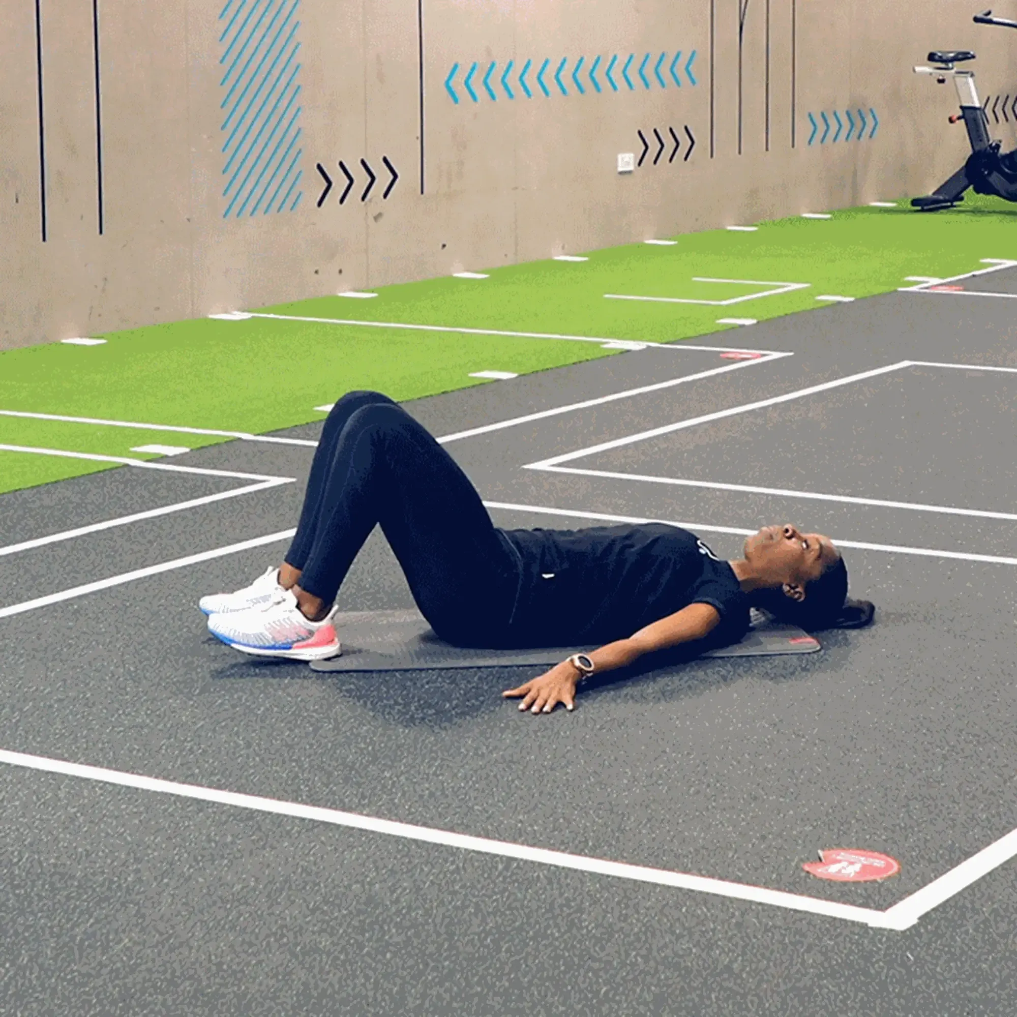 Woman lying on exercise mat with knees bent in a gym with marked flooring and bike