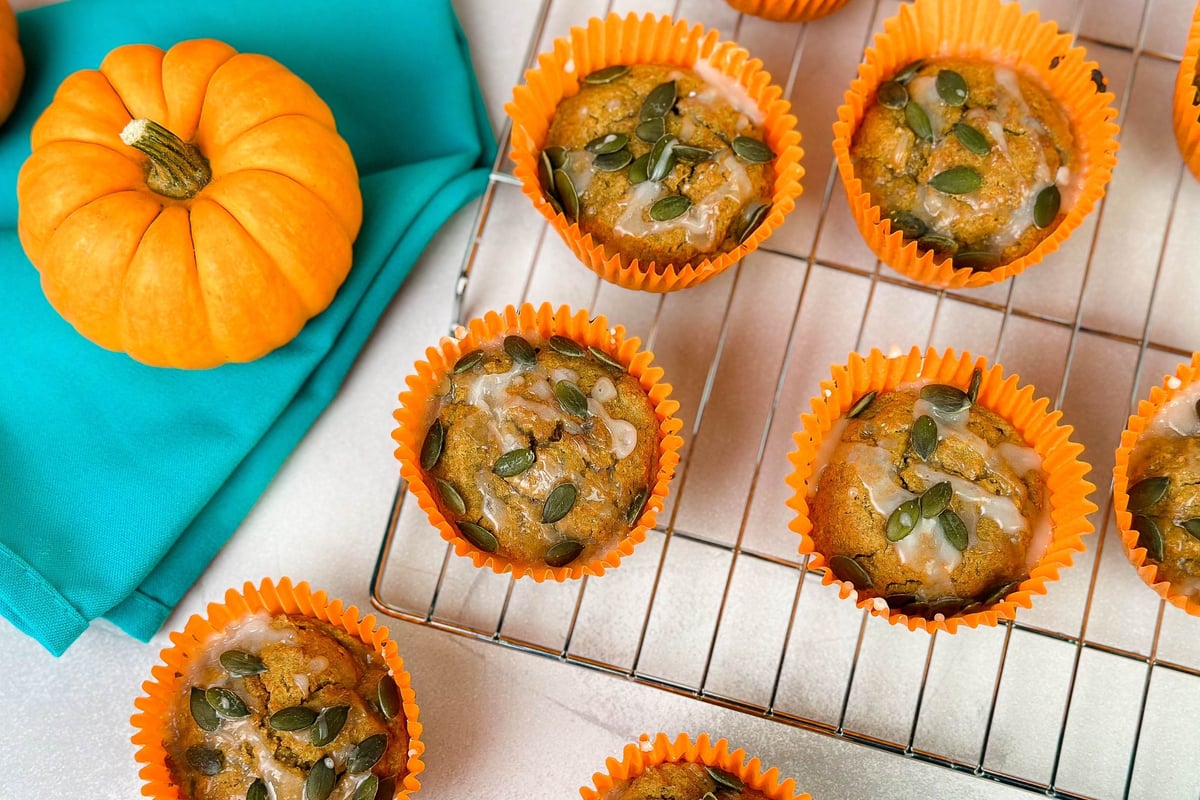 Pumpkin muffins topped with seeds in orange cases on a cooling rack beside small pumpkins