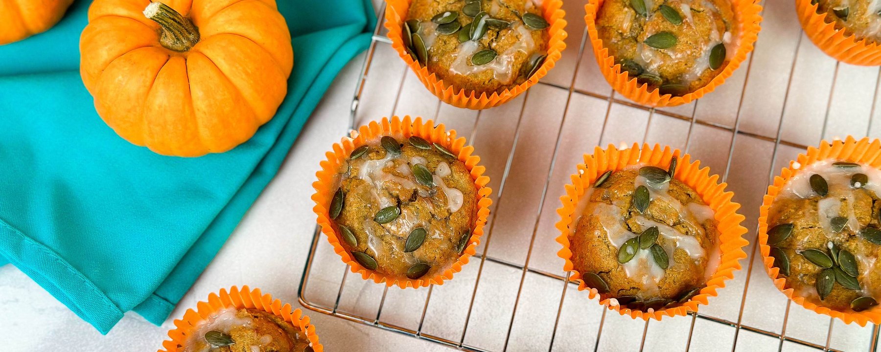 Pumpkin muffins topped with seeds in orange cases on a cooling rack beside small pumpkins