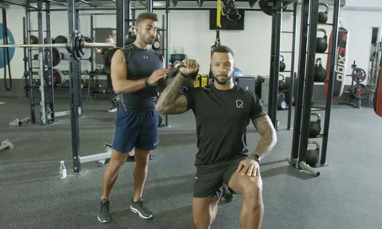 Man kneeling with kettlebell held overhead while trainer assists in gym