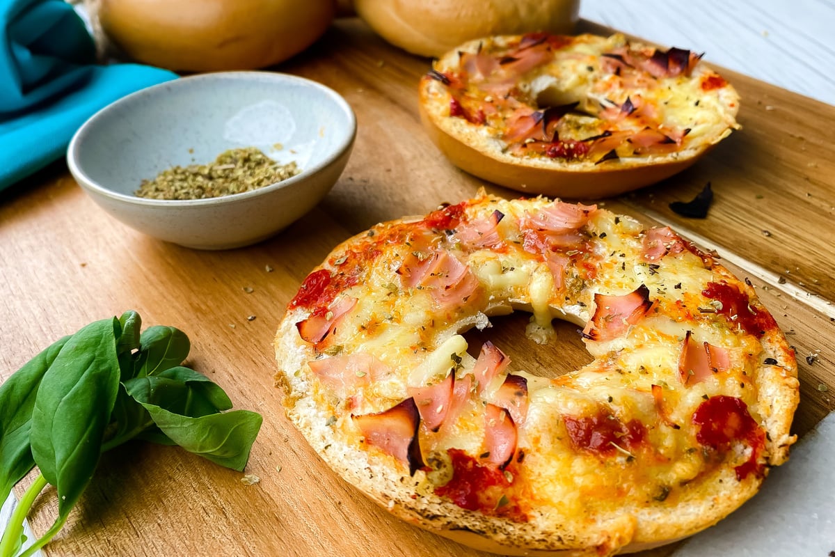 Two toasted bagel halves topped with melted cheese and ham on a wooden board