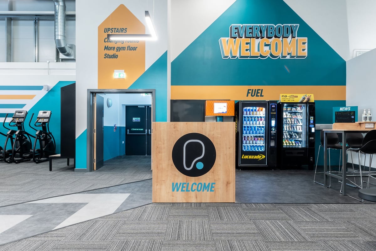 PureGym Kettering: Vending machines, snack racks, seating areas with "Everybody Welcome" signage indicating a gym lobby area.