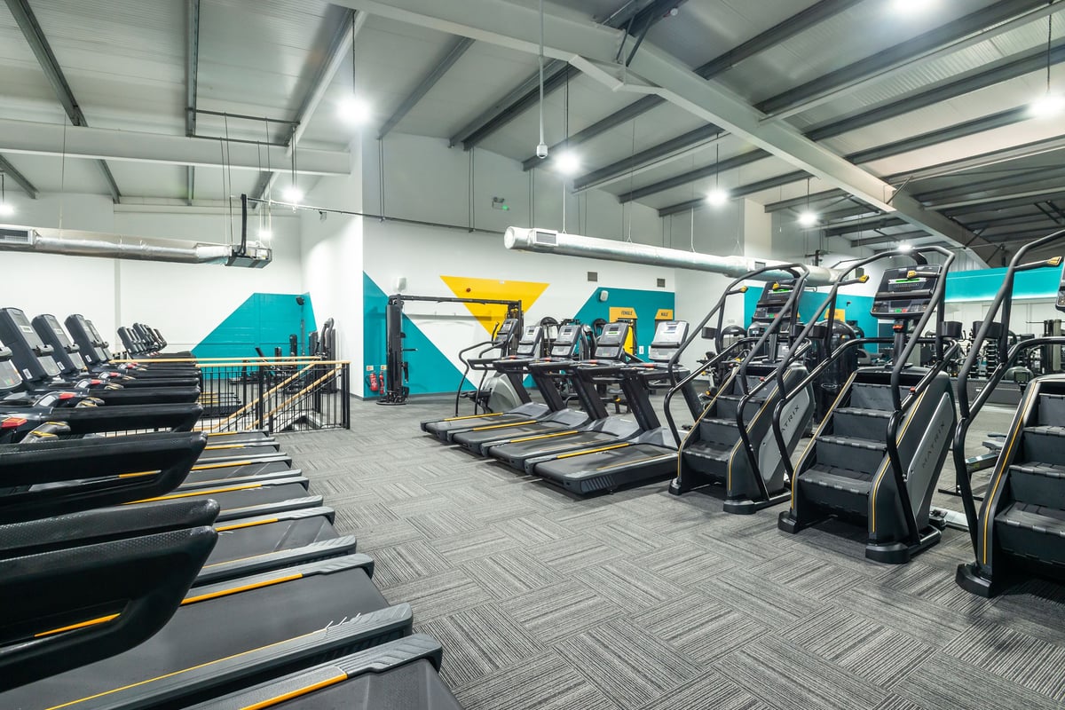 PureGym Wrexham: Rows of treadmills, ellipticals, weights benches and free weights in a brightly-lit gym space.