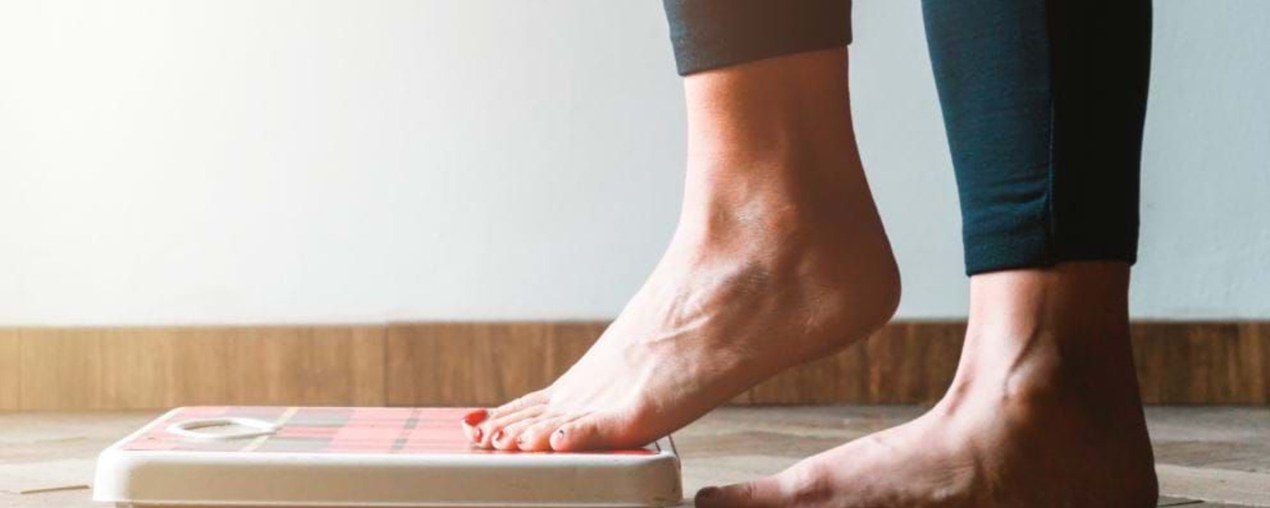 Bare feet stepping onto a weighing scale on a wooden floor against a white wall
