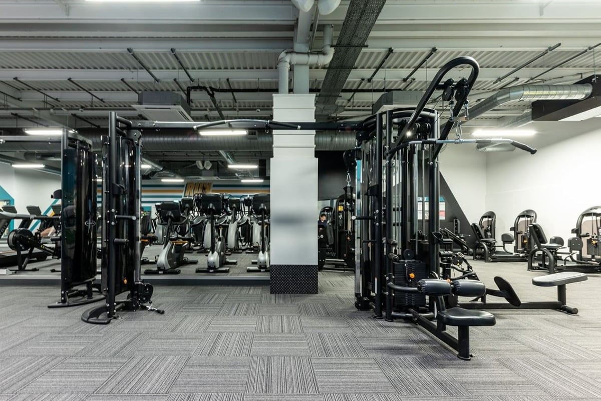 PureGym Cambridge Leisure Park: Rows of strength machines, weight racks, benches, and rubberized flooring in a dimly lit gym.