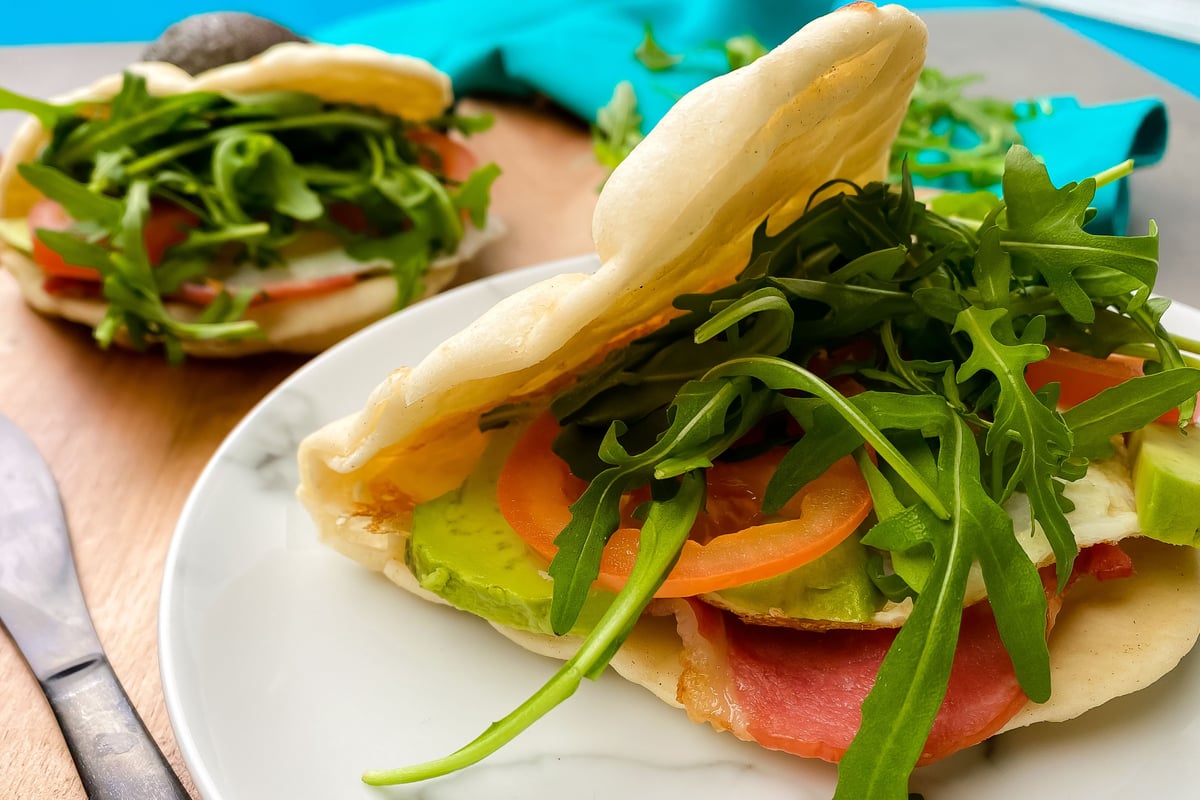 Flatbread filled with bacon, egg, rocket, avocado and sliced vegetables on white plate