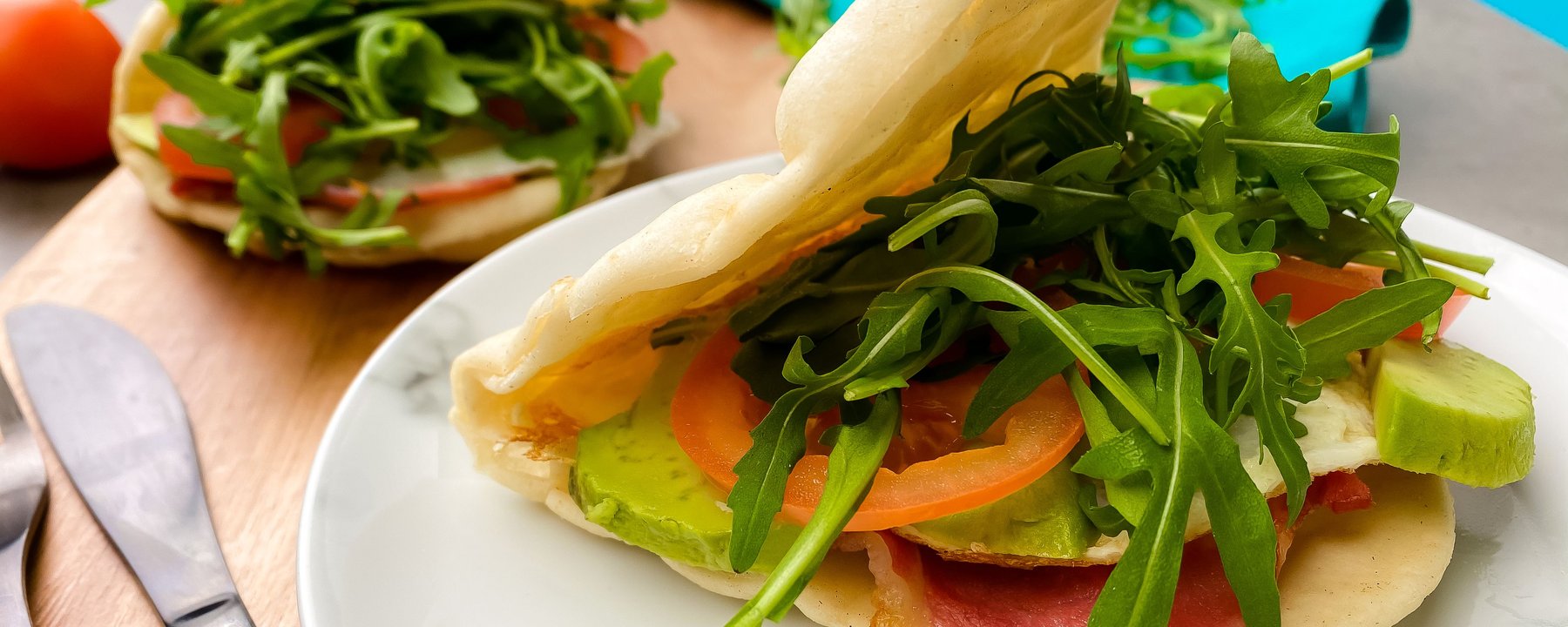 Flatbread filled with bacon, egg, rocket, avocado and sliced vegetables on white plate