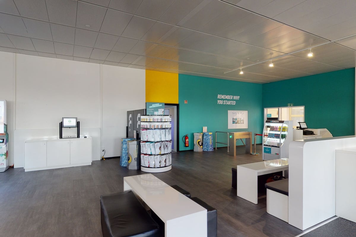 PureGym Aarau: Comptoir de fitness, étagères de rangement, coin salon avec tables, mur décoratif bleu sarcelle, parquet.