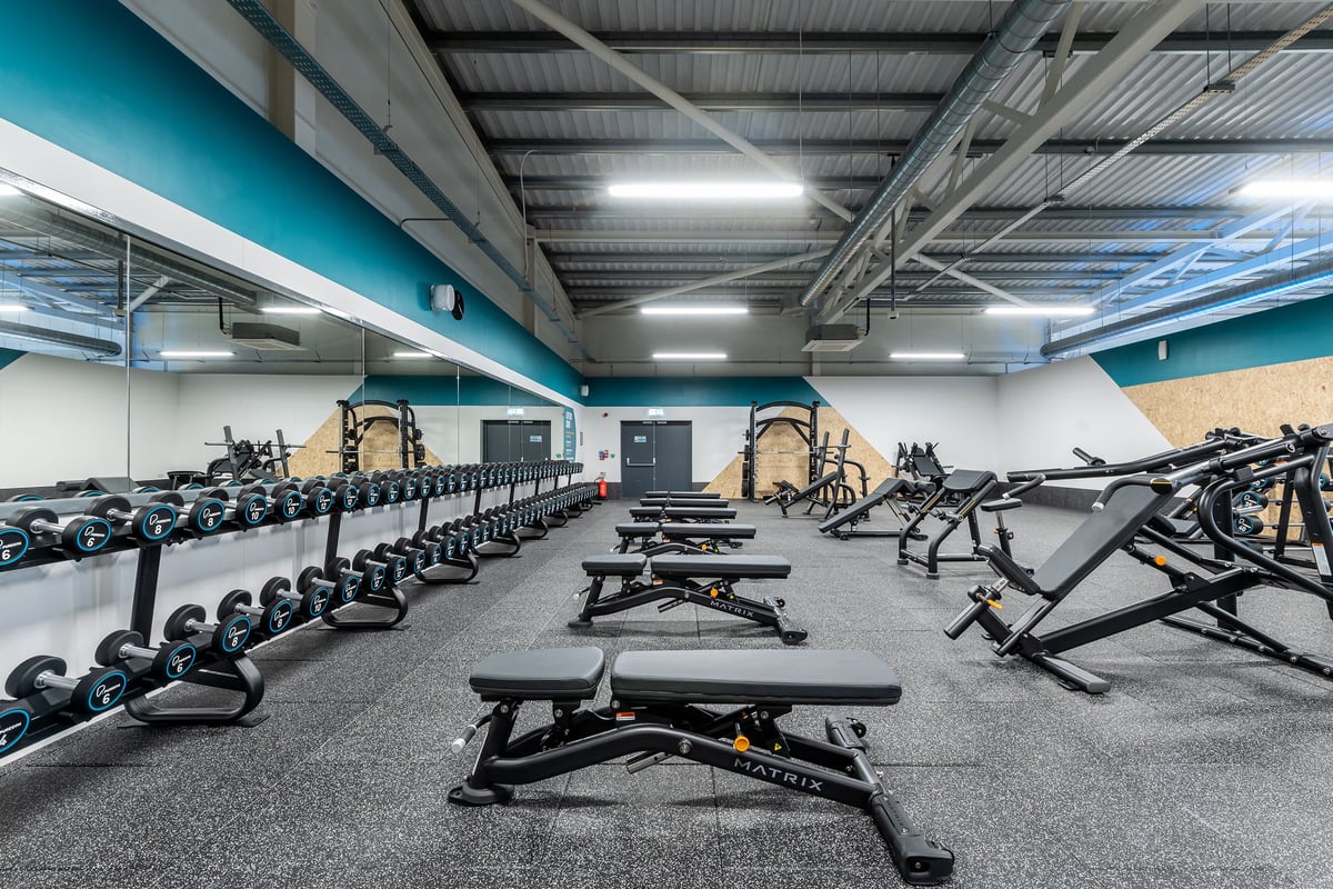 PureGym Barnsley: Rows of weight benches, squat racks, free weights, and rubberized flooring in a teal-colored gym.