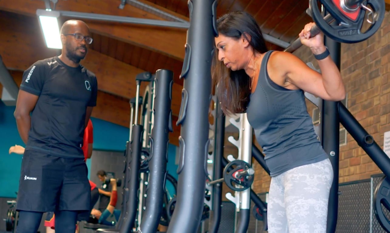 Woman lifting barbell on shoulders with trainer watching in gym setting