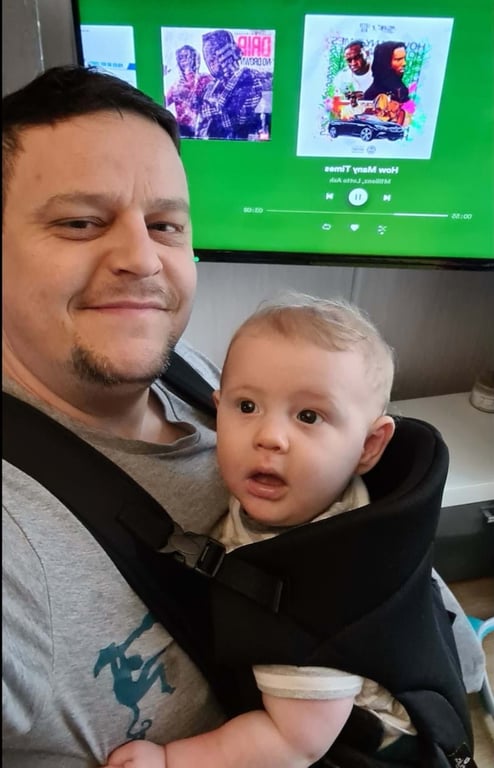 Man smiling with baby in carrier in front of a screen showing music covers and controls