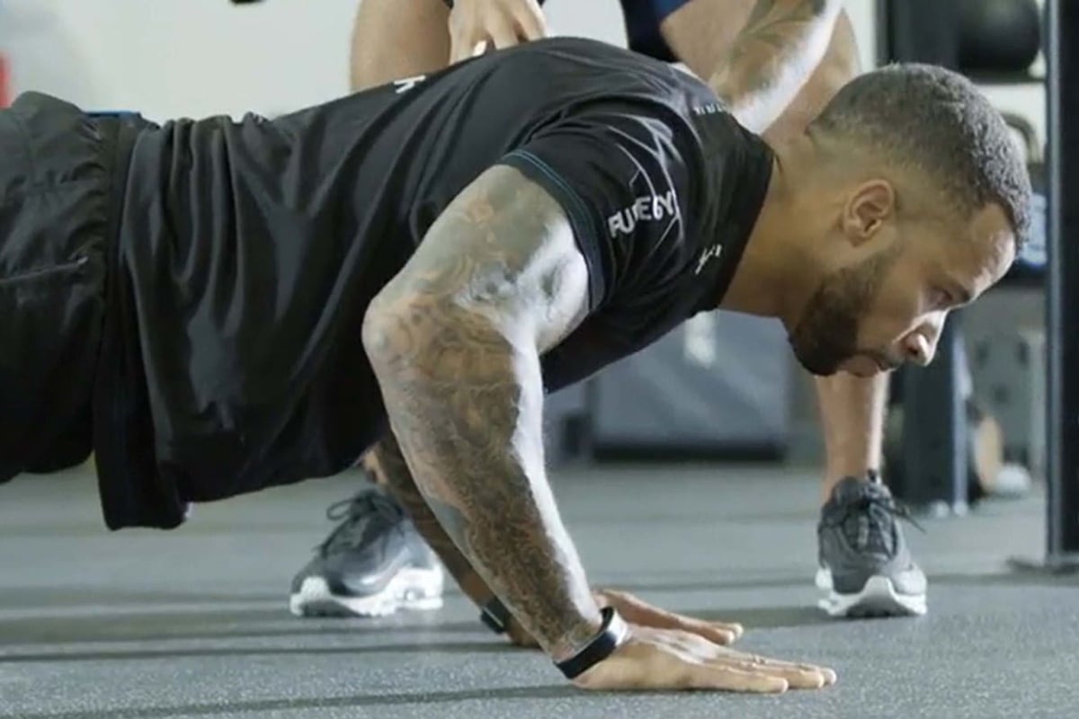 Man with tattoos doing a push-up in gym with another person standing behind him