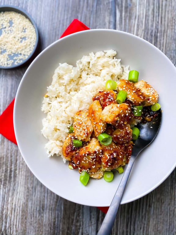 White rice with sesame chicken and chopped spring onions in a bowl with a spoon