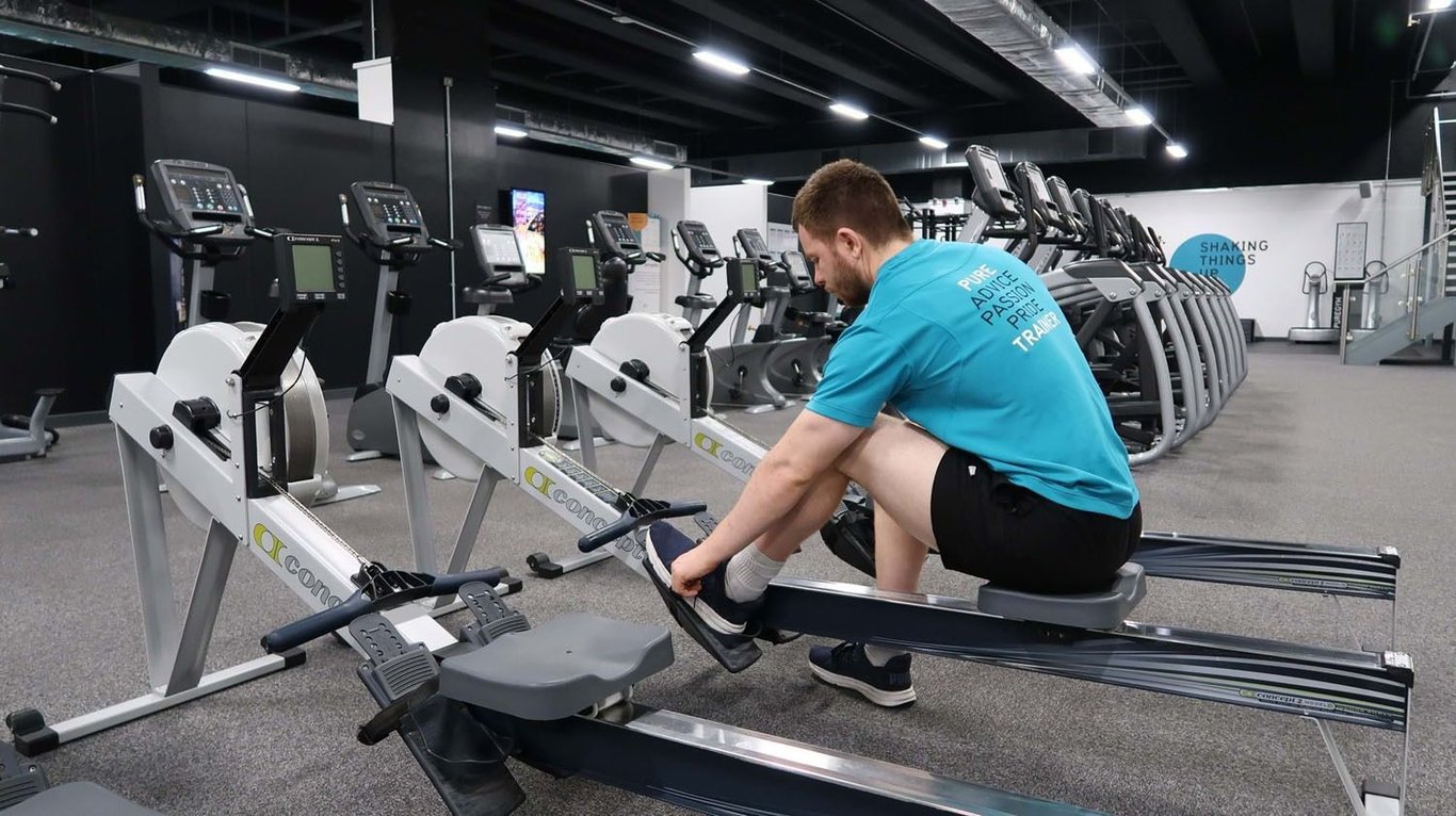 Man in blue shirt adjusting shoes on rowing machine in gym with cardio equipment
