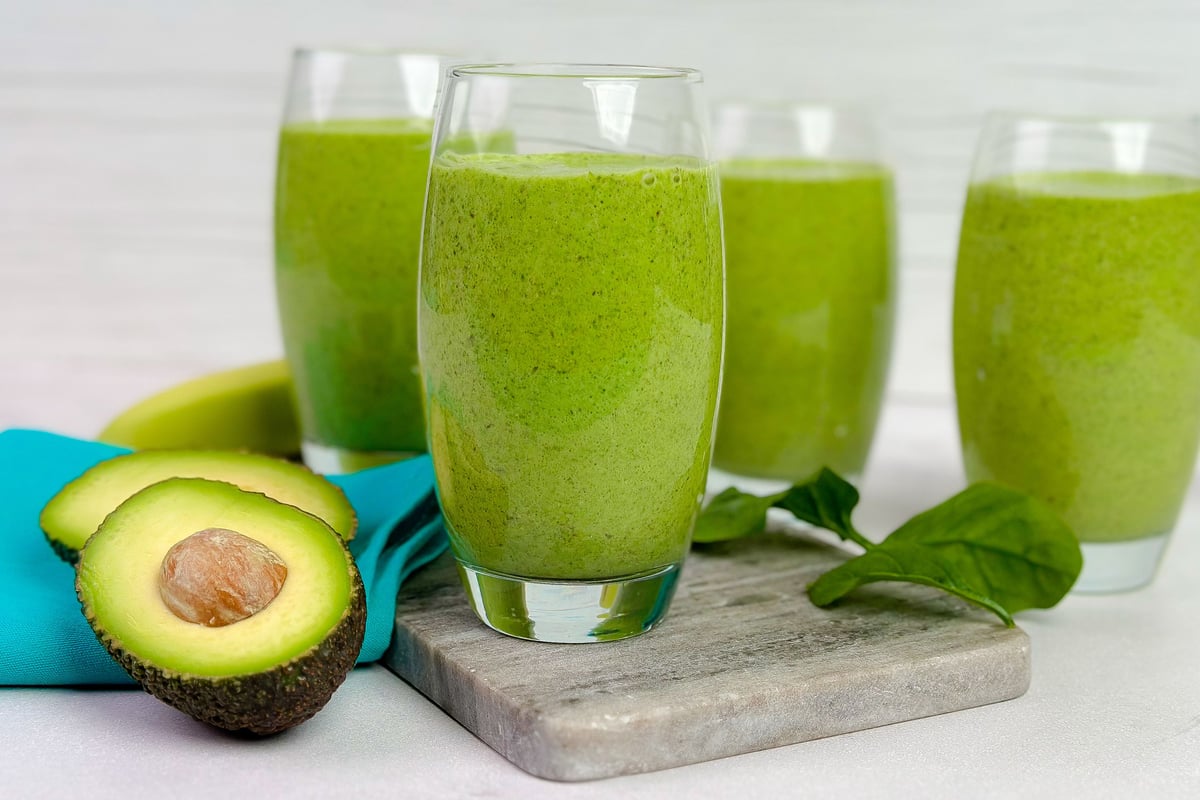 Four glasses of green smoothie on a wooden board with halved avocado and spinach leaves