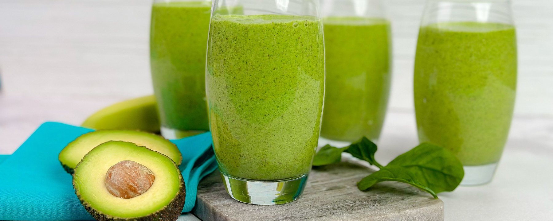 Four glasses of green smoothie on a wooden board with halved avocado and spinach leaves