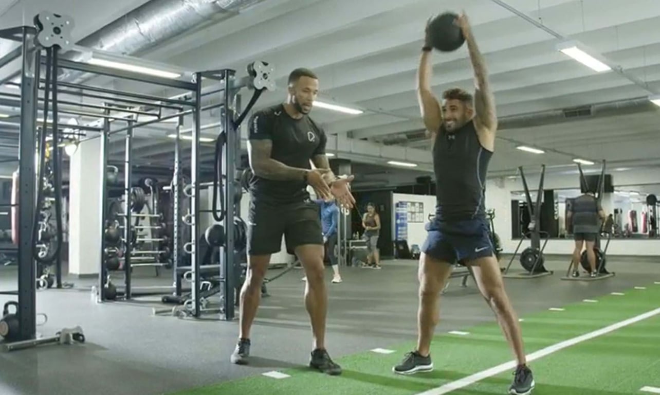 Man lifting medicine ball overhead while trainer watches in gym with equipment