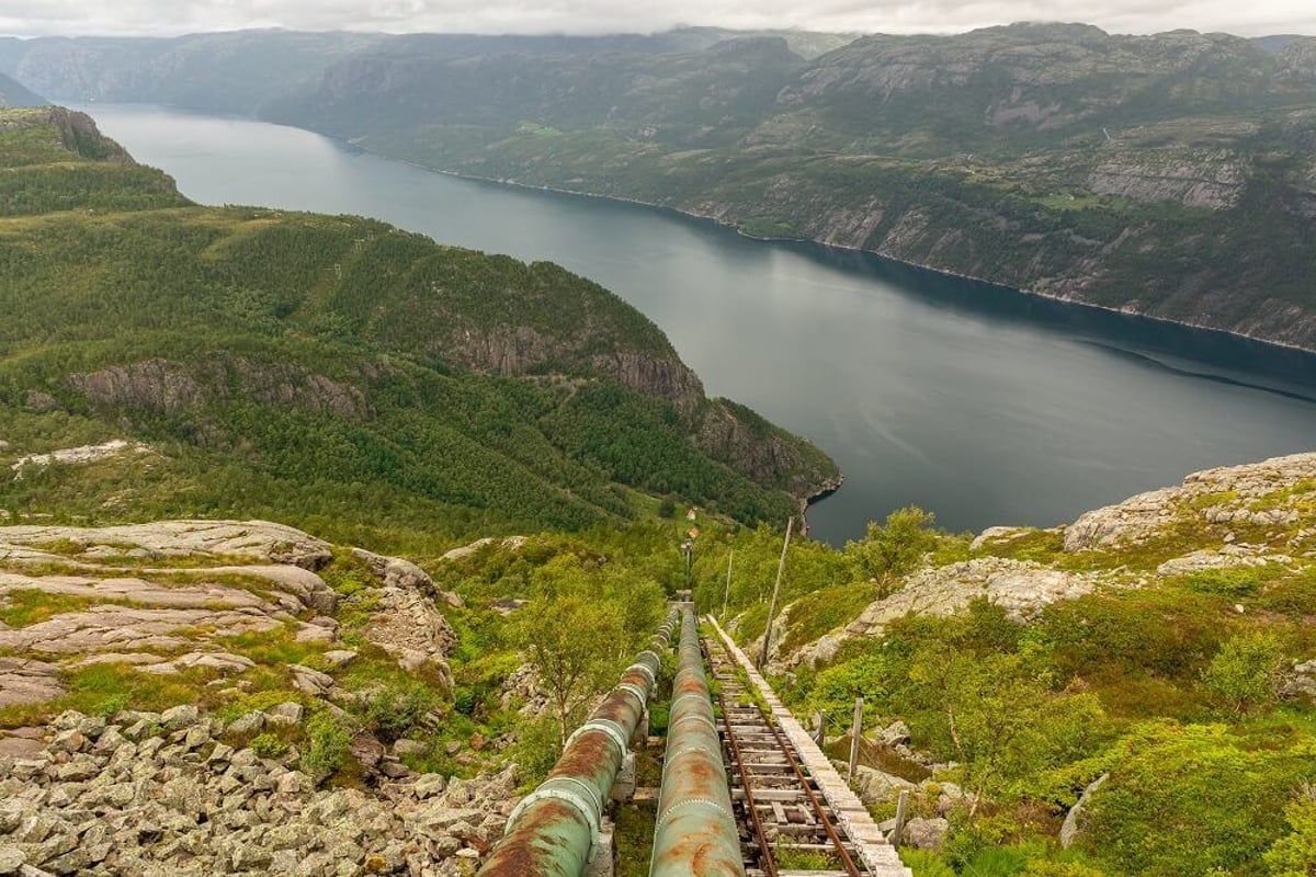 Rusty pipes and railway tracks descending a green hillside towards a fjord under cloudy sk