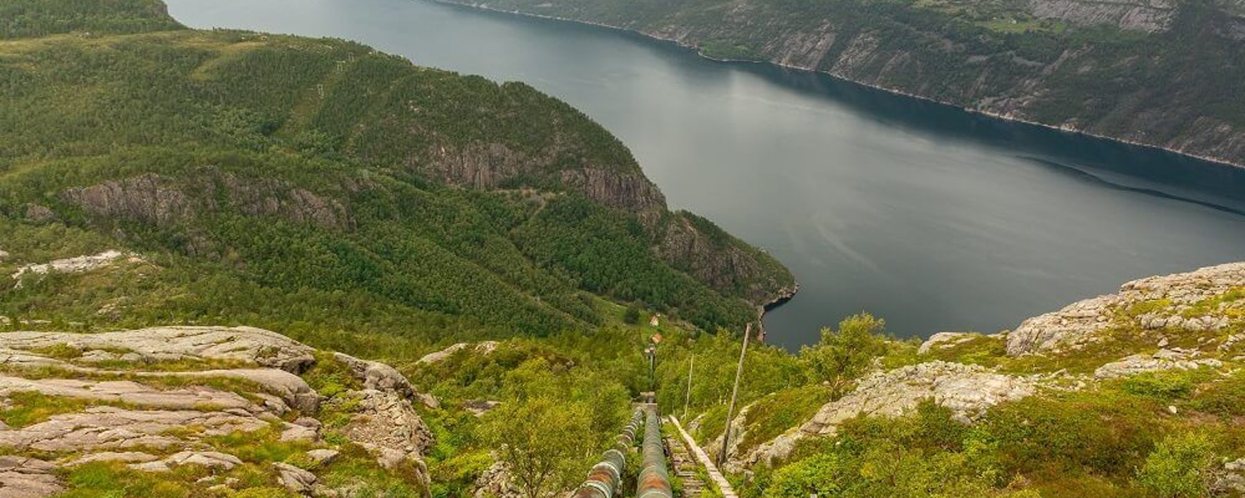 Rusty pipes and railway tracks descending a green hillside towards a fjord under cloudy sk