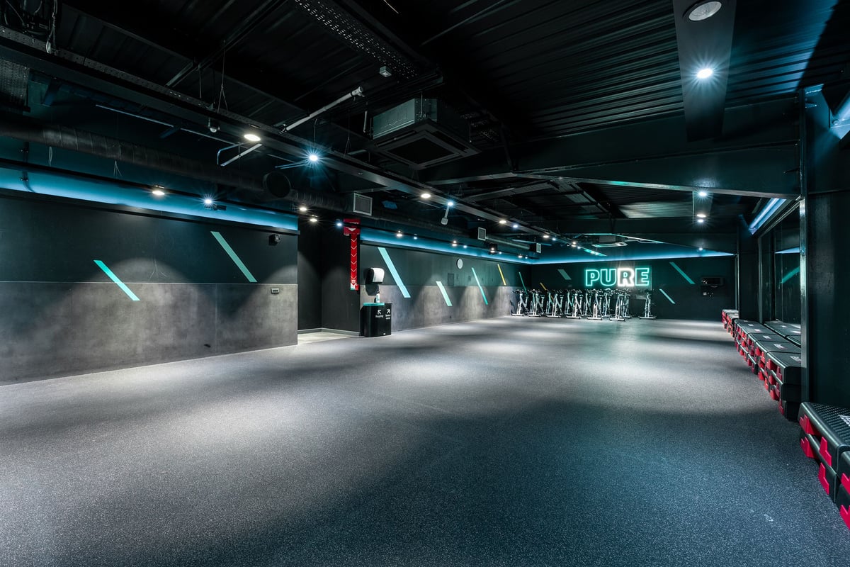 PureGym Leamington Spa: Empty studio with weight racks, rubberized flooring, and industrial lighting fixtures overhead.