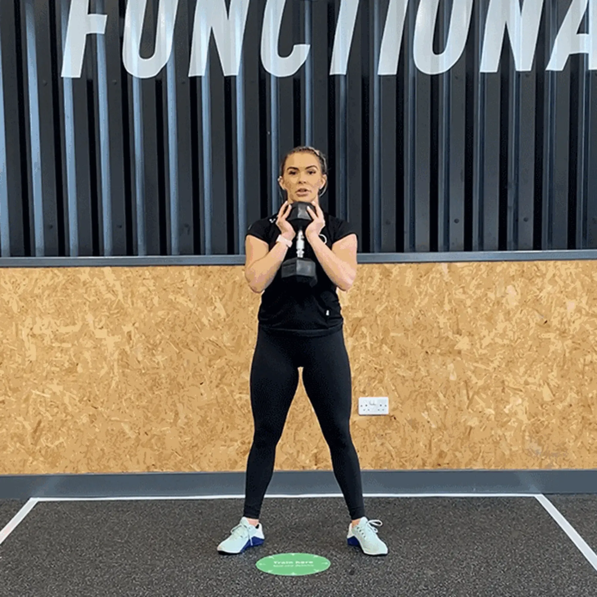 Woman in black workout clothes holding a dumbbell in front of her chest in a gym