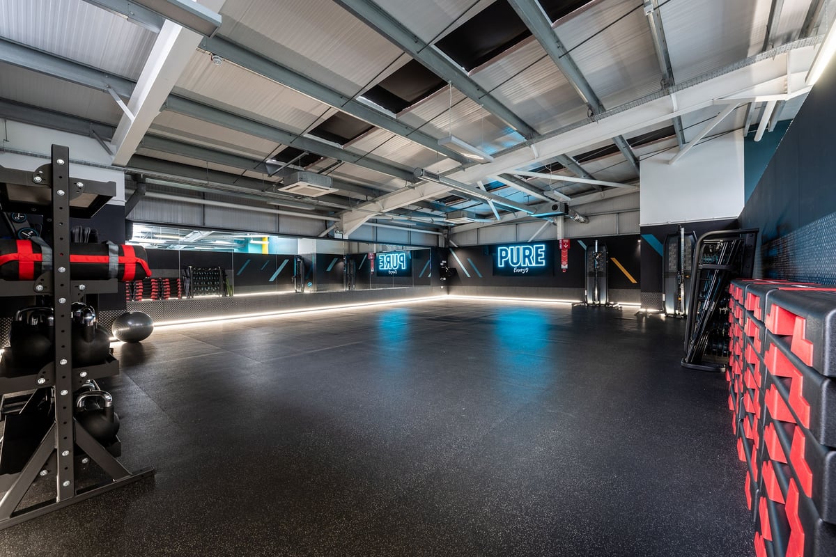 PureGym Wirral Bidston Moss: Mirrors, rubberized flooring, neon signage, and an industrial ceiling in a dark fitness space.