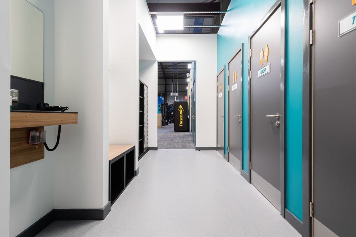 PureGym Elgin: Hallway with TV, wooden bench, framed artwork, and doors leading to fitness studios.