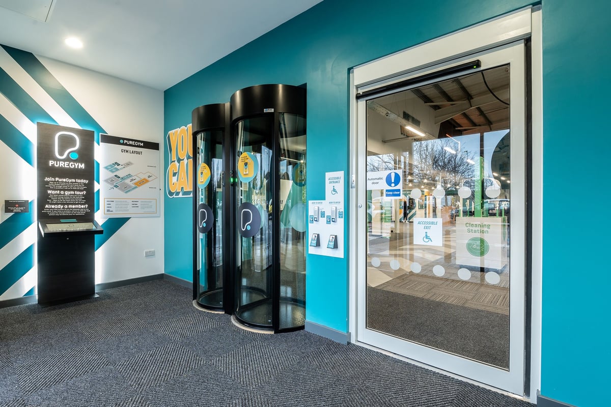 PureGym Ashford Warren Retail Park: Revolving security gates, teal walls, poster advertising facility capabilities.