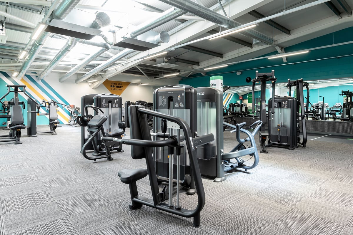 PureGym Colchester Retail Park: Rows of weight machines, benches, and rubberized flooring inside a gym with teal accents.