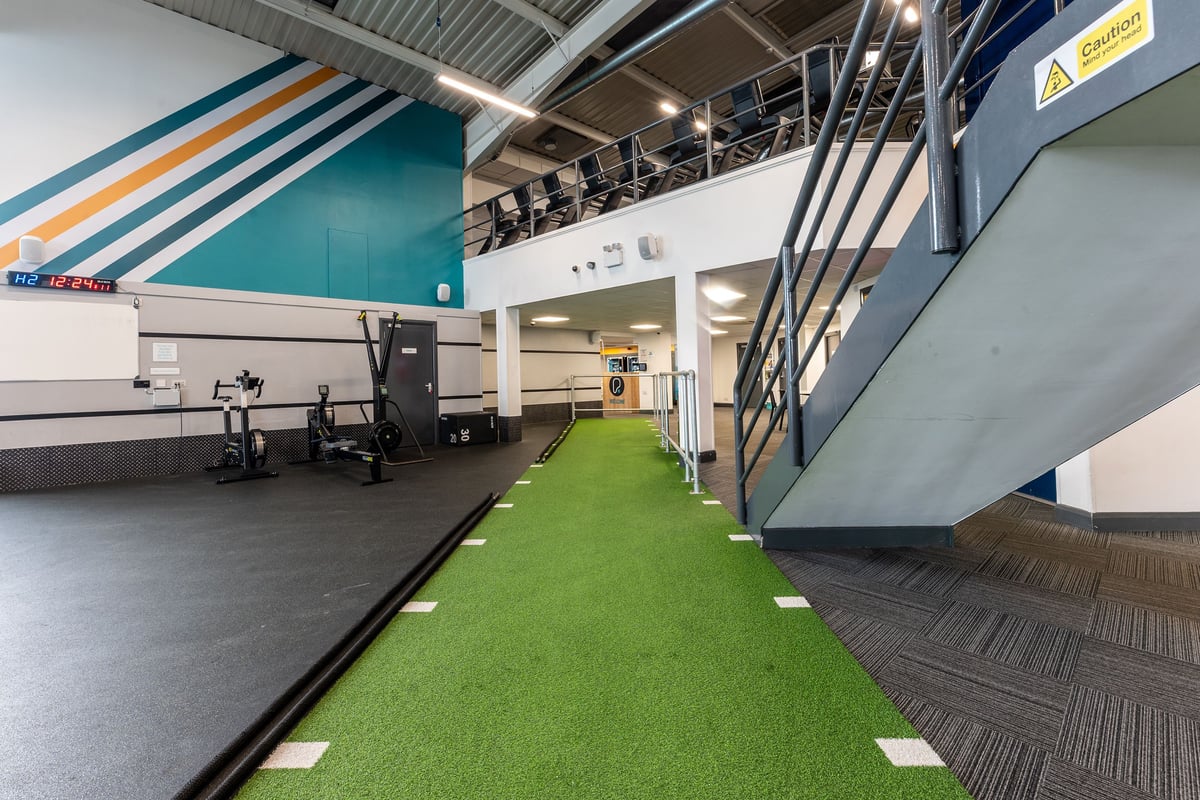 PureGym Brierley Hill: Padded astroturf with free weights and staircase, striped walls, industrial gym interior.