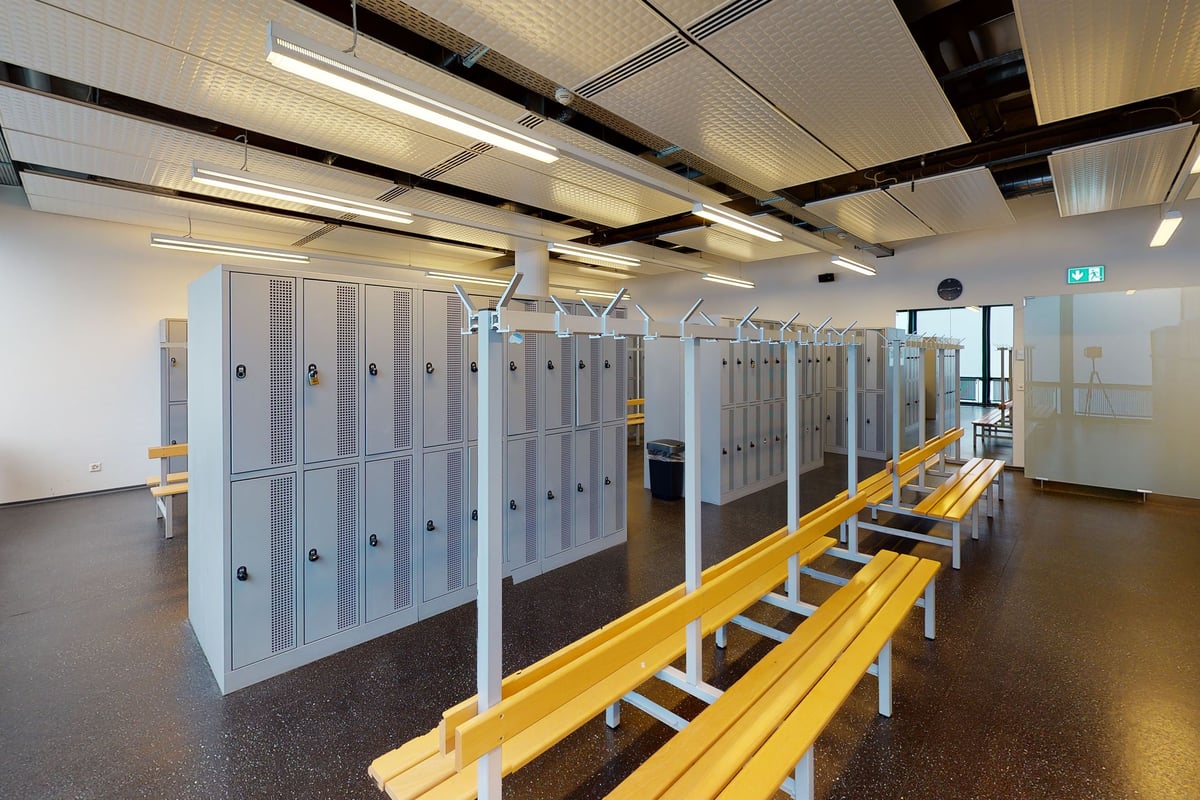 PureGym Biel Bienne: Casiers gris, bancs, balustrade jaune à l'intérieur des vestiaires d'un centre de remise en forme avec éclairage au plafond.