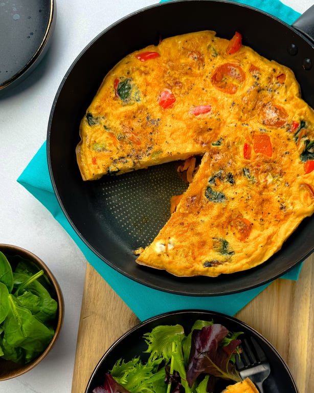 Vegetable omelette with a slice cut out in a frying pan on a blue cloth and wooden board