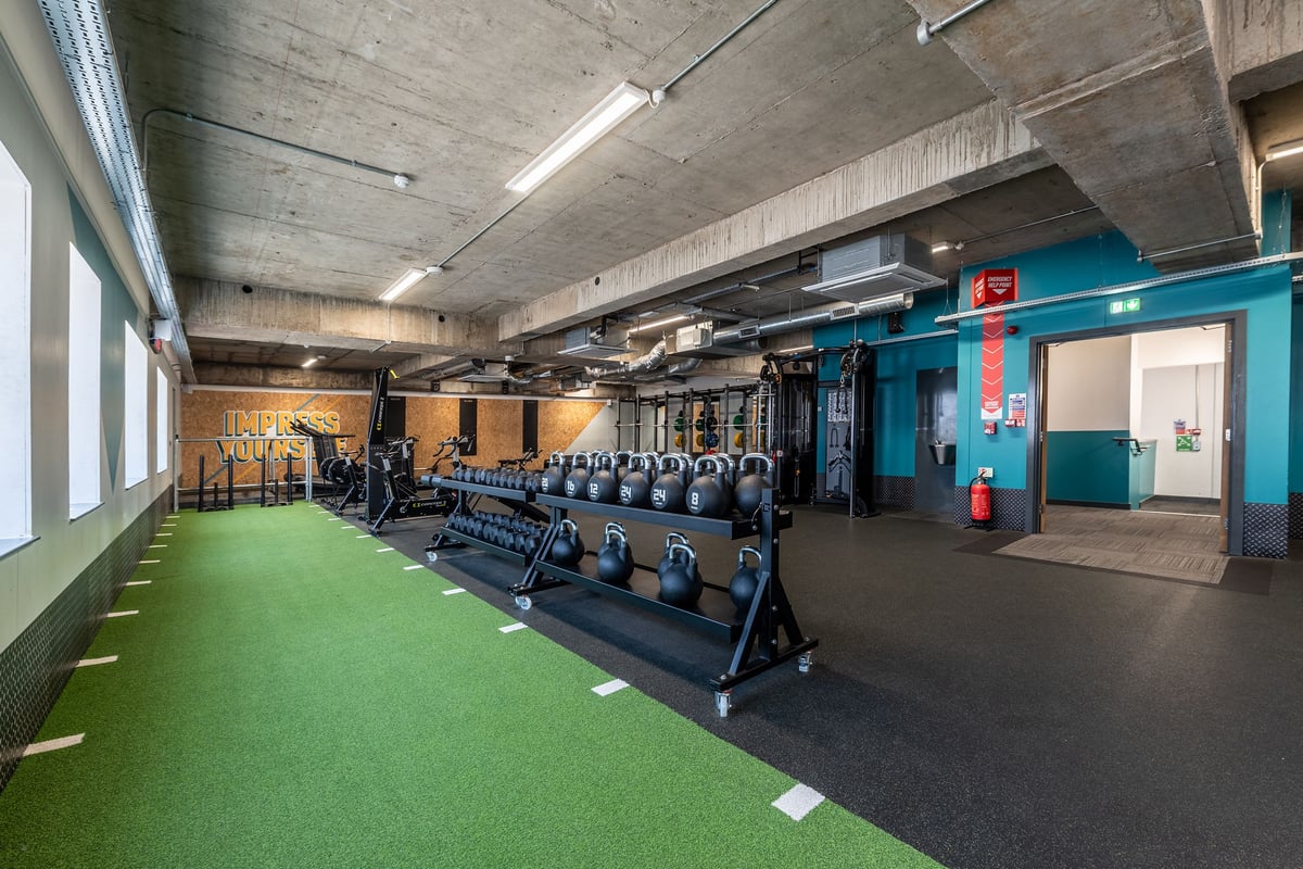 PureGym London Swiss Cottage: Rows of kettlebell racks, dumbbells, and rubberized turf flooring in a concrete-walled gym.