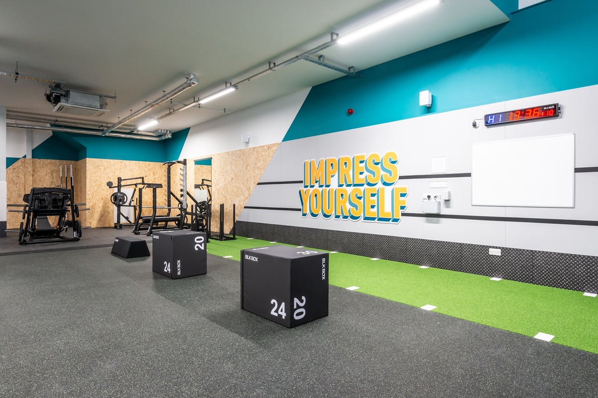 PureGym Market Harborough: Gym studio with lifting rigs, plyo boxes, weight plates, turf, and motivational wall branding.