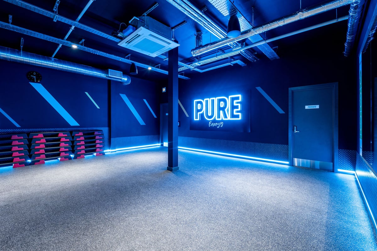 PureGym Cirencester: Weight benches, neon "Pure Energy" signage, industrial interior with blue lighting.