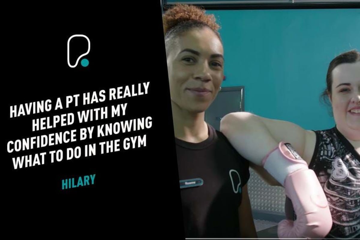 Two women smiling in a gym, one wearing pink boxing gloves and sportswear.