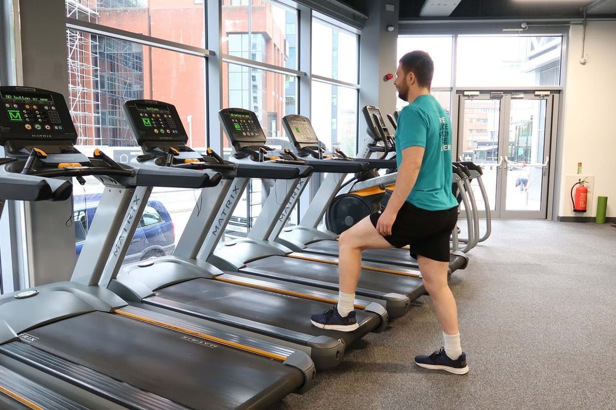 Man in sportswear stretching leg on treadmill in gym with large windows