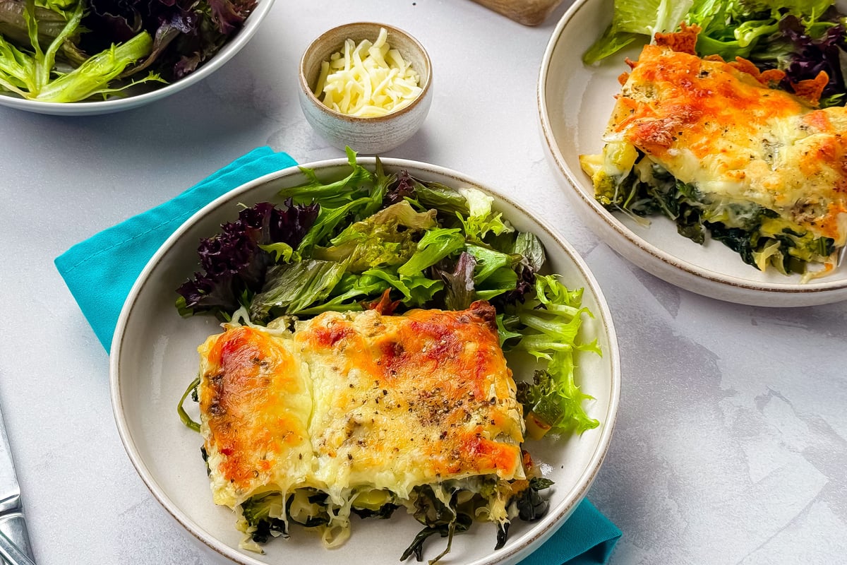 Two plates of baked spinach lasagne served with mixed salad leaves on a grey surface