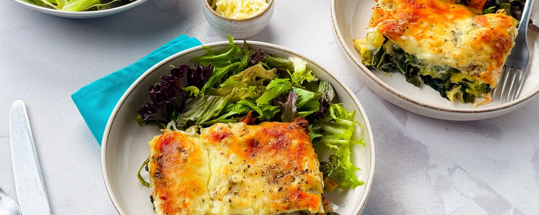 Two plates of baked spinach lasagne served with mixed salad leaves on a grey surface