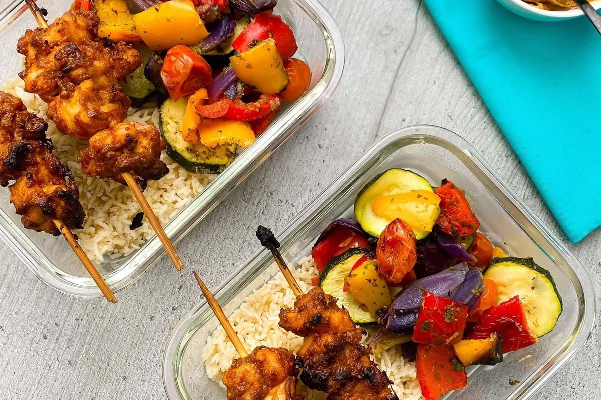 Two glass containers with grilled chicken skewers, rice, and mixed roasted vegetables.