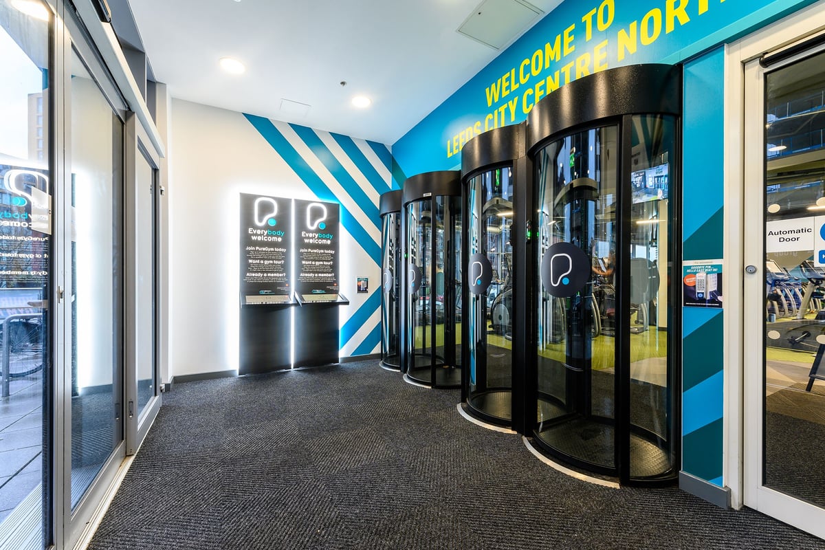 PureGym Leeds City Centre North: Entrance lobby with branded signage, glass doors, and digital kiosk displays.