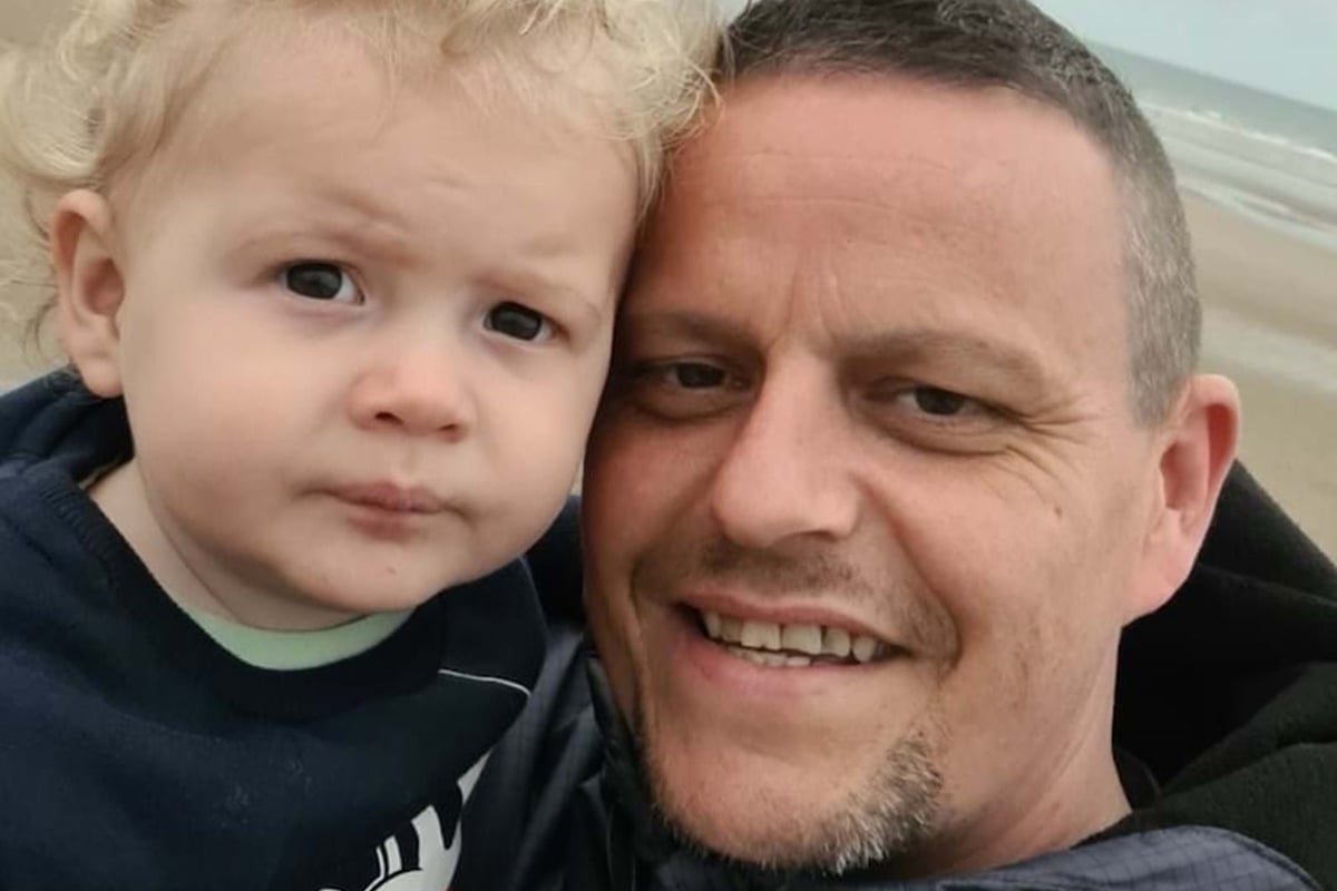 Man smiling holding a toddler with curly blonde hair on a beach background