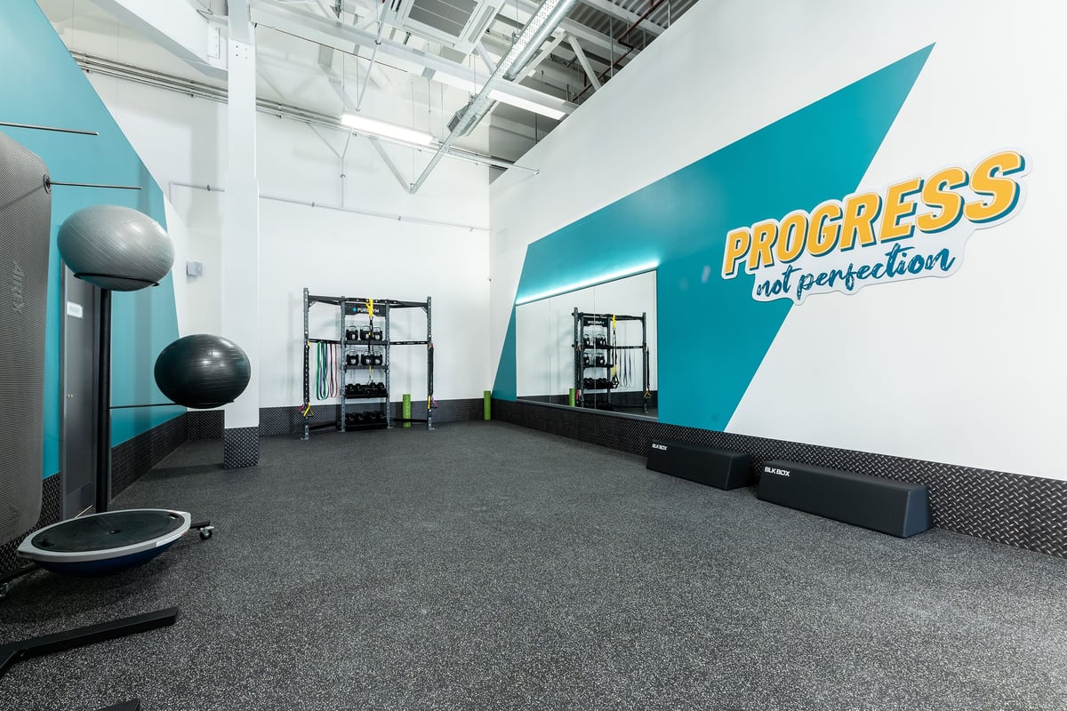 PureGym Rushden: Gym studio with exercise balls, free weights, rubberized flooring, teal accent wall with "Progress" logo.